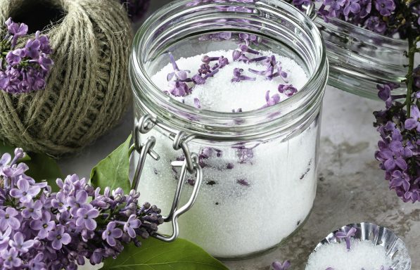 A jar of cream with purple flower petals, surrounded by lilac blossoms and twine.